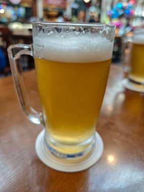 Canlı bir bar ortamında, koyu ahşap bir masanın üzerinde kremalı köpüklü bir bardak soğuk bira duruyor. Parlak renkler ve rahat bir atmosfer birliktelik anlarını davet eder.