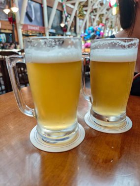 Üstünde bol köpük olan iki buzlu bira bardağı şenlikli bir ortamda ahşap bir masada dinleniyor. Arka planda renkli balonlar ve Noel süslemeleri arkadaşlarla kutlamak için neşeli ve rahat bir atmosfer yaratır..