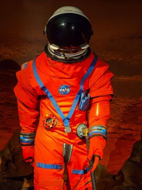 Parlak turuncu uzay giysisi içinde NASA logosunun olduğu bir astronot resmi. Arka plan, kayaları ve kırmızımsı tonu ile Mars yüzeyini anımsatıyor ve ilginç bir keşif atmosferi yaratıyor. Detaylı kıyafet gelişmiş teknolojiyi yansıtıyor.