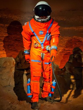 Turuncu bir uzay giysisi içindeki bir astronot Mars arazisinde adım atarak uzay keşfi arayışını vurguluyor. Sahne, dünya dışı bir çevrede cesaret ve keşif duyguları uyandırıyor..