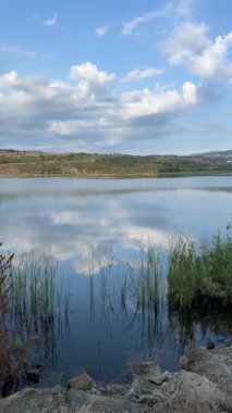 Sakin bir göl, yumuşak bulutları ve mavi gökyüzünü yansıtır. Sahil boyunca sazlıklar ve uzakta yumuşak tepeler vardır. Bir cep telefonu videosundan elde edilen huzurlu bir doğal manzara yaratır.