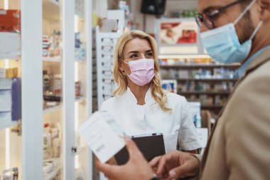Young business man choosing and buying drugs in a drugstore while talking with attractive female pharmacist. She helping him with expert advices. They are wearing protective face masks against virus infection. clipart