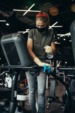 Genç bayan işçi temizlik yapıyor ve pahalı spor aletlerini alkol spreyi ve bezle dezenfekte ediyor. Coronavirus küresel dünya salgını ve sağlık koruma önlemleri.