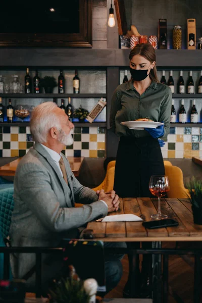 The waitress serves a delicious meal to the senior businessman at the restaurant. She wears a protective mask as part of security measures against the Coronavirus pandemic.