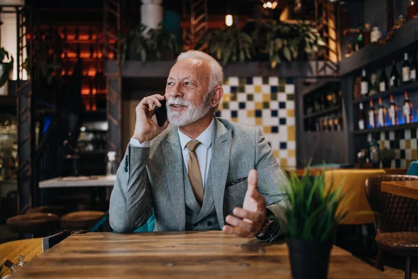 Happy businessman sitting in restaurant and waiting for lunch. He is using smart phone and talking with someone. Business seniors lifestyle concept.
