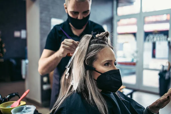 美容師は女性の髪を染めており 彼のクライアントに箔で髪のハイライトを作ります 彼女はウイルスのパンデミックに対する保護として保護面マスクを着用している — ストック写真