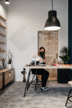 Stüdyosunda ya da atölyesinde çalışan kağıt tasarımcısı ve sanatçı. Virüs salgınına karşı koruyucu yüz maskesi takıyor..