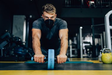 Modern spor salonunda egzersiz yapan koruyucu maskeli yakışıklı bir erkek sporcu. Güçlü gölgeleri olan karanlık, çamurlu bir ışık. Salgın spor konsepti.