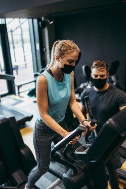 Modern spor salonunda vücut çalışmasında genç ve çekici bir kadın kişisel fitness hocası ya da koçuyla birlikte. Mesafelerini koruyorlar ve koruyucu yüz maskeleri takıyorlar. Coronavirus dünya salgını ve spor teması.