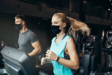 Modern spor salonunda koşu bandında koşan genç, zinde bir kadın ve erkek. Mesafelerini koruyorlar ve koruyucu yüz maskeleri takıyorlar. Coronavirus dünya salgını ve spor teması.