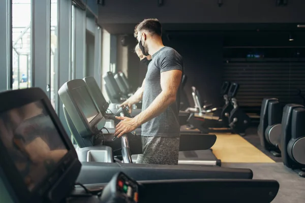 stock image Young fit woman and man running on treadmill in modern fitness gym. They keeping distance and wearing protective face masks. Coronavirus world pandemic and sport theme.