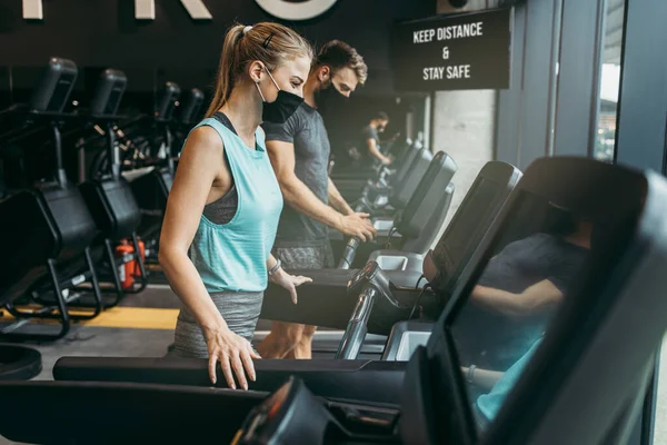 Young fit woman and man running on treadmill in modern fitness gym. They keeping distance and wearing protective face masks. Coronavirus world pandemic and sport theme.