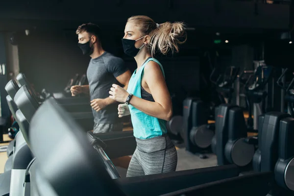 Young fit woman and man running on treadmill in modern fitness gym. They keeping distance and wearing protective face masks. Coronavirus world pandemic and sport theme.