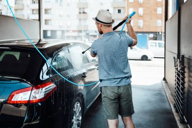 Gündüz vakti araba yıkama istasyonunda yüksek basınçlı su kullanan şapkalı genç bir adam arabasını yıkıyor..