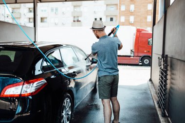 Gündüz vakti araba yıkama istasyonunda yüksek basınçlı su kullanan şapkalı genç bir adam arabasını yıkıyor..