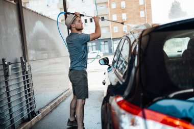 Gündüz vakti araba yıkama istasyonunda yüksek basınçlı su kullanan şapkalı genç bir adam arabasını yıkıyor..