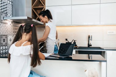 Covid-19 salgını sırasında ya da karantina sırasında e-öğrenim seansı yapan küçük bir kız. Anne mutfakta çalışıyor ve arka planda öğle yemeği hazırlıyor.. 