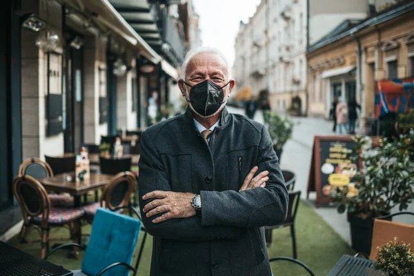 Confident senior businessman standing in front of exclusive restaurant. He is wearing protective N-95 face mask as protection against virus infection. Empty street in background. Coronavirus senior people lifestyle concept.