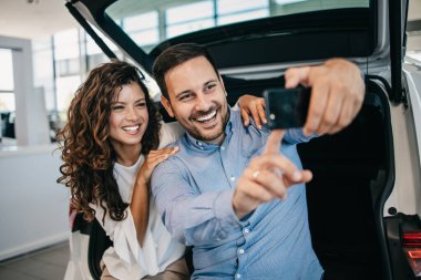 Mutlu orta yaşlı çift, galeride yeni araba seçerken ve alırken eğleniyor. Yeni arabalarıyla selfie çekiyorlar..