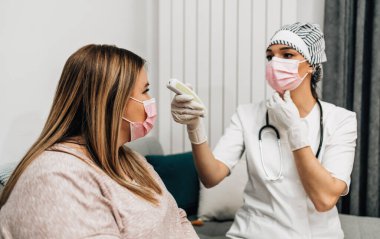 Orta yaşlı hasta bir kadın kanepede otururken kadın doktor vücut ısısını kızılötesi alın termometresi tabancasıyla kontrol ediyor..