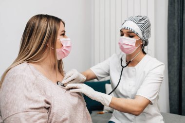 Genç bayan doktor ev ziyareti sırasında hastasını steteskopla kontrol ediyor. Kanepede oturan hasta kadın Coronavirus veya Covid-19 virüs enfeksiyonundan muzdarip.. 