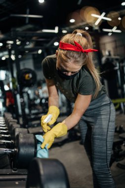 Genç bayan işçi temizlik yapıyor ve pahalı spor aletlerini alkol spreyi ve bezle dezenfekte ediyor. Coronavirus küresel dünya salgını ve sağlık koruma önlemleri.
