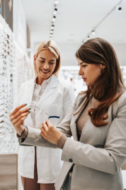 Modern optik mağazada gözlük çerçevesi seçen güzel ve modaya uygun bir kadın. Kadın satıcı uzmanı doğru karar vermesine yardımcı oluyor..