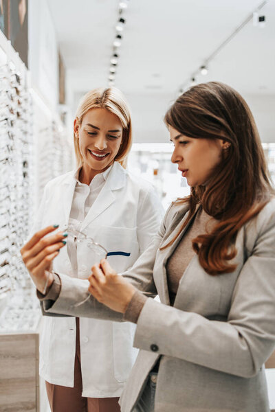 Beautiful and fashionable woman choosing eyeglasses frame in modern optical store. Female seller specialist helps her to make right decision.