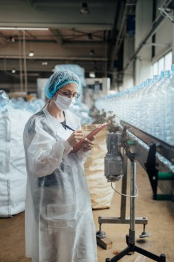 Tıbbi malzeme araştırma ve üretim fabrikasında çalışan ve sevkiyattan önce damıtılmış su kutularını kontrol eden koruyucu yüz maskeli bir kadın işçi. Denetim kalite denetimi. 