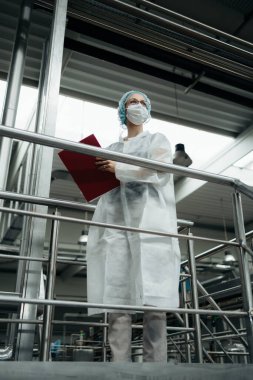 Koruyucu iş kıyafetlerinde çalışan, tıbbi malzeme araştırma ve üretim fabrikasında çalışan kadın işçi. Kontrol köprüsünde duruyor ve üretim hattını izliyor. Denetim kalite denetimi.