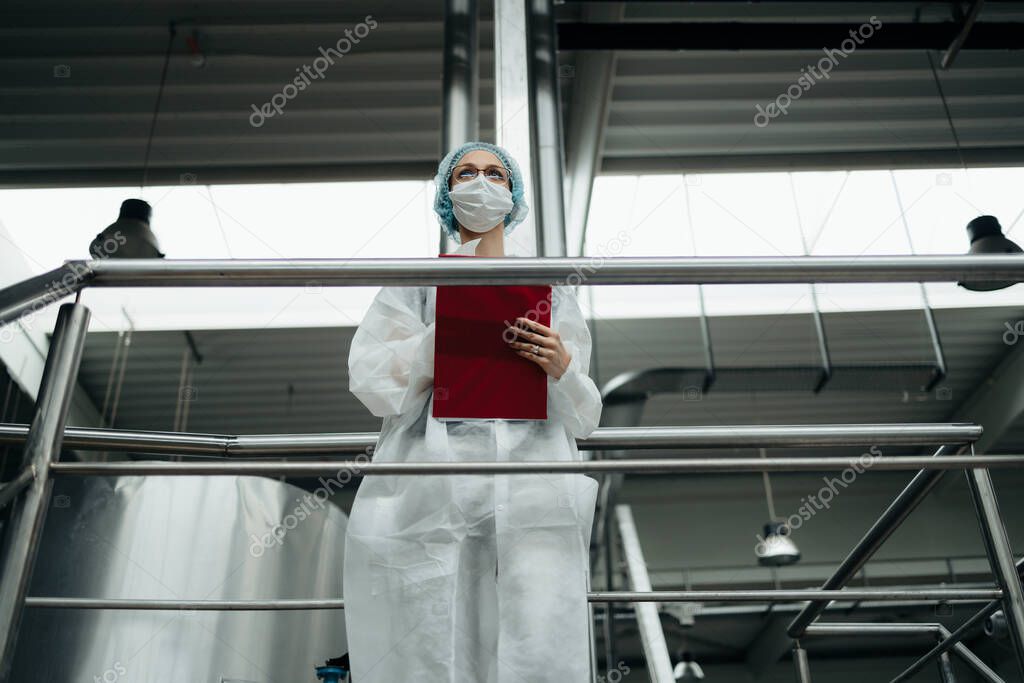 Female worker in protective workwear working in medical supplies research and production factory. She is standing on control bridge and watching at production line. Inspection quality control.
