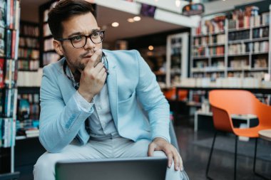 Genç çekici bir adam modern kitapçıda oturuyor, dizüstü bilgisayarını okuyor ve kullanıyor..