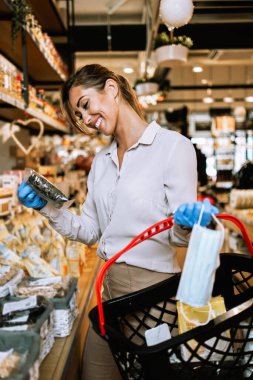Yüzü koruyucu maskeli ve eldivenli genç ve zarif bir kadın modern bir süpermarkette ya da markette sağlıklı yiyecek ve içecek satın alıyor. Salgın ya da salgın yaşam tarzı ve tüketim kavramı.