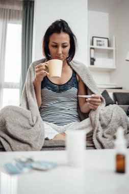 Grip hastası ve yorgun orta yaşlı bir kadın. Evdeki kanepede oturuyor, termometreyi tutuyor ve bej battaniyeye sarılı vücut ısısını ölçüyor..