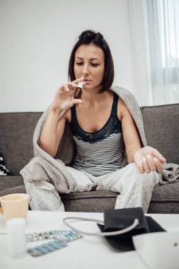 Grip hastası ve yorgun orta yaşlı bir kadın. Evdeki kanepede oturuyor, termometreyi tutuyor ve bej battaniyeye sarılı vücut ısısını ölçüyor..