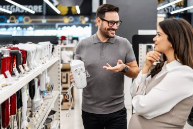 Neşeli bir elektronik mağazasında çeşitli ürünler sergilerken hamile bir müşteriye ev aletleri seçmesinde yardımcı olan gülümseyen mağaza asistanı
