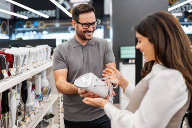 Neşeli bir elektronik mağazasında çeşitli ürünler sergilerken hamile bir müşteriye ev aletleri seçmesinde yardımcı olan gülümseyen mağaza asistanı