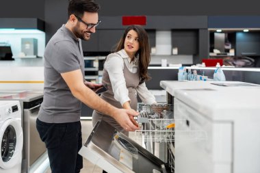 Elinde tablet olan gülümseyen bir satıcı hamile bir müşteriye bulaşıkçıları gösteriyor ve tartışıyordu. Modern bir ev aletleri mağazasından en iyi seçenekleri seçiyordu.