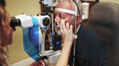 Üst düzey bir erkek hastanın gözlerini modern kliniğinde bir yarık lambasıyla inceleyen kadın göz doktoru görüşünü dikkatlice kontrol ediyor ve olası göz hastalıklarını teşhis ediyor.