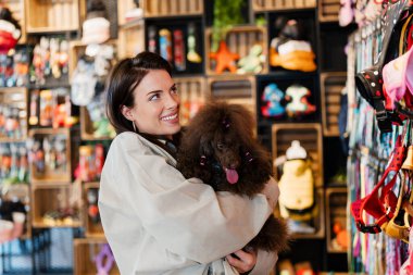 Bir perakende evcil hayvan dükkanında renkli evcil hayvan koşum takımlarına ve malzemelere bakarken gülümseyen ve kaniş köpeğini tutan bir kadın, hayvan sahipliğini ve bakımını kavramsallaştırıyor.