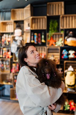 Bir perakende evcil hayvan dükkanında renkli evcil hayvan koşum takımlarına ve malzemelere bakarken gülümseyen ve kaniş köpeğini tutan bir kadın, hayvan sahipliğini ve bakımını kavramsallaştırıyor.