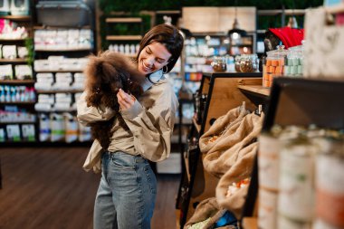Şirin kahverengi bir kaniş tutan ve modern bir evcil hayvan dükkanında yüksek kaliteli evcil hayvan mamaları seçen bir kadın, insan-hayvan bağını ve hayvan sahipliğini resmediyor.