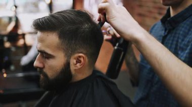 Berber, müşterinin saçını saç spreyiyle dikkatlice şekillendiriyor. Modern bir berber dükkanında sakallı ve solgun saçlı bir adama profesyonel bakım hizmeti veriyor.