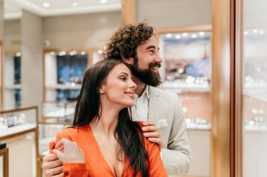 Genç çift, lüks bir butik vitrininde saatlere ve takılara hayranlıkla bakarken birbirlerine yakın duruyorlar. Romantik vitrinlere bakıyorlar ve özel bir gün için hediyeler seçiyorlar.