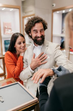 Gülümseyen mutlu bir adam lüks bir kol saatini denerken satış asistanı bunu ayarlıyor ve ortağı zarif bir kuyumcuda şık, kendine güvenen bir satın alma deneyimi yaşıyor.