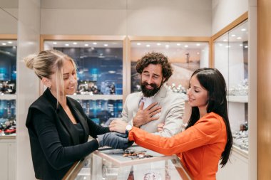 Satıcı kadın, gülümseyen bir çifte modern bir butikte lüks bir kol saatini denemesinde yardımcı olur. Özel bir hediye seçerler, özenli müşteri hizmetlerinden ve zarif ortamlardan hoşlanırlar.