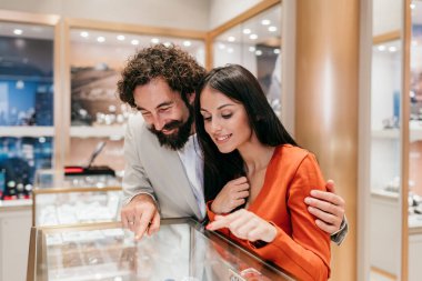 Genç çift, sofistike bir kuyumcuda pahalı mücevherlere sevinçle bakıyor, vitrindeki eşyalara bakıyor, tüketim ve lüks alışverişleri gösteriyor.