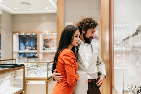 Zarif bir butikte lüks saat ve takılara hayran olan şık bir çift romantik, lüks bir alışveriş gününde vitrin alışverişi ve hediye seçiyor.