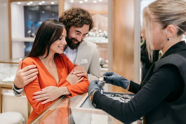 Genç zarif çift lüks kuyumcuda lüks kol saatini deniyor. Gülümsüyorlar ve dikkatli satış yardımı ve zarif atmosferi olan mükemmel bir saat seçiyorlar.