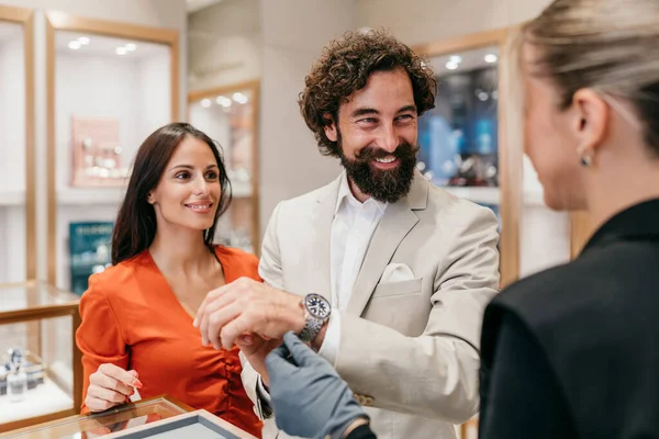 Gülümseyen mutlu bir adam lüks bir kol saatini denerken satış asistanı bunu ayarlıyor ve ortağı zarif bir kuyumcuda şık, kendine güvenen bir satın alma deneyimi yaşıyor.